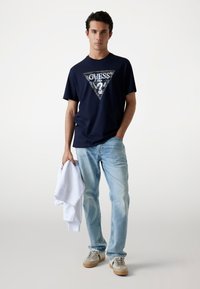 Marineblaue Baumwoll-T-Shirt mit einem dreieckigen silbernen Grafiklogo, kombiniert mit hellblauen Jeans und einem weißen Sweatshirt in der Hand. Beigefarbene Sneakers.