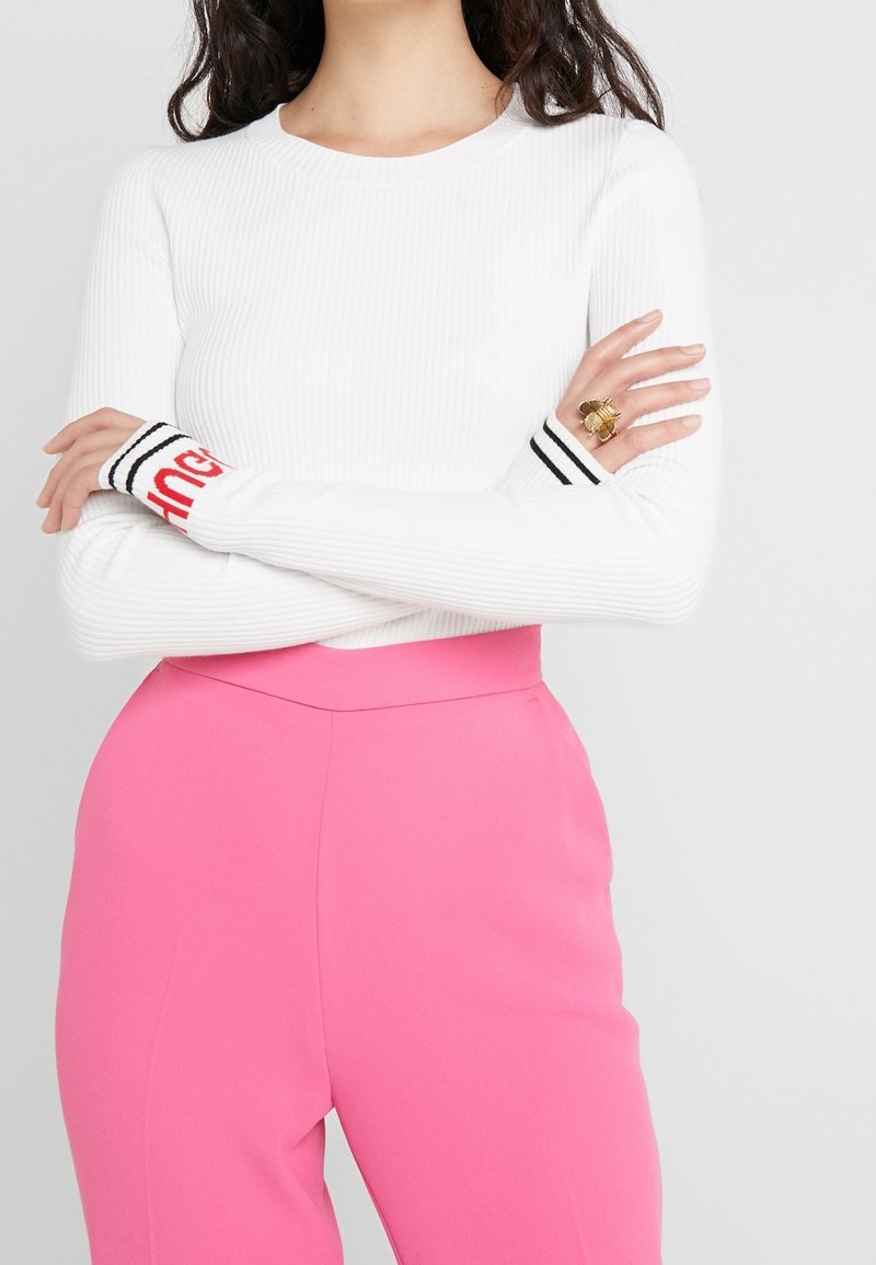 Top bianco a maniche lunghe con coste, con polsini a strisce nere e rosse, abbinato a pantaloni rosa a vita alta. Dettagli notevoli includono un anello decorativo.