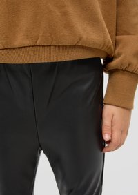 Bruine gemeleerde trui met geribbelde mouwen, gecombineerd met strakke zwarte neplederen broek. Hand zichtbaar, rustend op de broek.