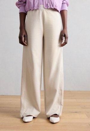 Osoba nosząca kremowe spodnie o szerokich nogawkach, białe spiczaste buty i liliową krótką kurtkę, stojąca na drewnianej podłodze.