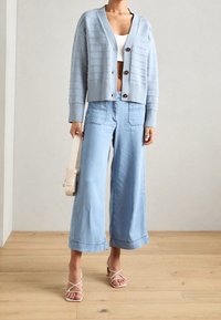 Žena nosí zkrácený světle modrý cardigan, bílý zkrácený top, široké světle džínov�é kalhoty, béžové sandály na podpatku a drží béžovou kabelku.