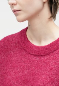 Zbliżenie dolnej części twarzy i szyi osoby noszącej teksturowany ciemnoróżowy sweter z prążkowanym kołnierzem na białym tle.