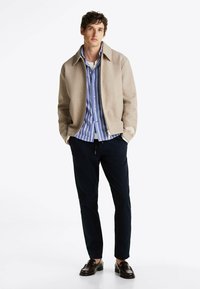 Giacca beige a trama, camicia a righe blu e bianche, pantaloni blu navy, mocassini marroni, con una vestibilità moderna e design con colletto.