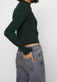 Top de manga larga de color verde oscuro con textura acanalada, combinado con vaqueros de denim gris de cintura alta, que cuentan con bolsillos frontales y traseros y un diseño ajustado.