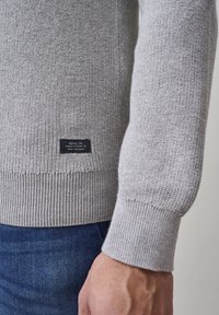 Sivi pleteni pulover s rebrastim rubom i manžetama. Ima malu crnu oznaku na dnu s bijelim tekstom. Dizajniran za uski fit.