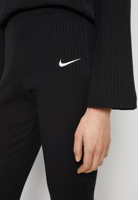 Melni ribētie legingi ar gludu tekstūru un baltu Nike logo akcentu sānos. Tuvplāns uz roku, kas atspiedusies pret audumu.