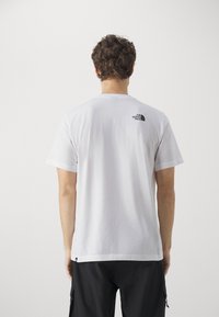 Camiseta de algodón blanca con mangas cortas, que presenta un pequeño logo negro en la parte superior de la espalda y un dobladillo recto. La tela parece suave.