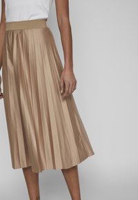 Jupe midi plissée de couleur beige clair, fabriquée en tissu lisse et brillant. Elle dispose d'une large ceinture élastique pour le confort.