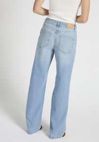 Osoba u svijetloplavim trapericama ravnih nogavica i bijelom majicom bez rukava stoji ispred jednobojne bijele pozadine.