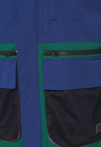 Blå och grön jacka med dragkedjefickor, mesh-detaljer och strukturerad tyg. Innehåller ytterligare accentfärger och sömnadsdetaljer.