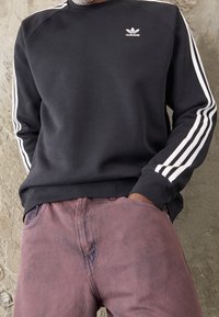 Man i svart Adidas-tröja med vita ränder på ärmarna och blek lila byxor, stående mot en texturerad vägg.