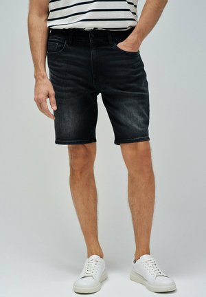 Pantalones cortos de mezclilla oscura de corte ajustado, con un diseño clásico de cinco bolsillos, detalles cosidos y cierre con botón. Combinados con zapatillas blancas.