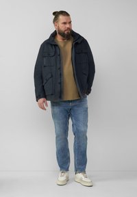 Marineblauwe jas met capuchon, meerdere voorzakken, over een tan shirt, gecombineerd met lichtblauwe jeans en witte sneakers met beige accenten.
