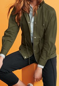 Olijf groene jas met een koordkap, voorrits en zijzakken, laag over een lichtblauw shirt, gecombineerd met donkere broek.