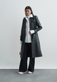 Black leather trench coat with button front, dual patch pockets, and wide lapels, layered over a white shirt and black wide-leg pants.
