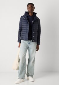 Marineblå puffet jakke med lynlås og trykknaplukning, med en quiltet tekstur, kombineret med lyseblå wide-leg jeans og hvide sneakers.