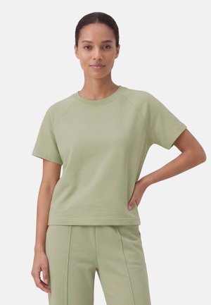 Donna con capelli legati che indossa una camicia verde chiaro a maniche corte e pantaloni coordinati, in piedi con una mano sul fianco.