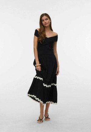 Nő fekete vállvillantós felsőben és hozzá illő midi szoknyában, fehér hullámos szegéllyel, fekete szandált és bézs karkötőket viselve, fehér háttér előtt áll.