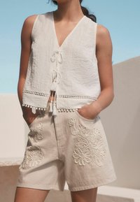 Crop top en lin blanc avec un col en V et des détails à nouer. Shorts beiges ornés de broderies florales et de poches latérales. Accents de franges occasionnels.