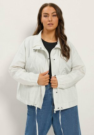 Femme aux longs cheveux bruns portant une veste gris clair à carreaux sur un haut noir et un jean bleu, ajustant la fermeture avant de la veste.