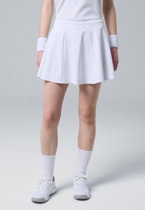 Persona che indossa una gonna plissettata bianca, scarpe da ginnastica bianche, calzini bianchi e polsini bianchi su uno sfondo grigio chiaro uniforme.