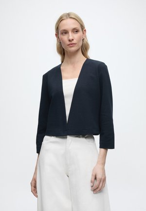 Frau mit langen blonden Haaren, trägt eine schwarze, offene Jacke über einem weißen Oberteil und weißen Hosen, steht vor einem einfarbigen Hintergrund.