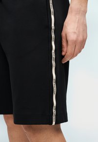 Juodi medvilniniai šortai su šoniniu tinklelio intarpu ir smėlio spalvos firminiu juosteliu. Paprasto dizaino, ilgis žemiau kelių.