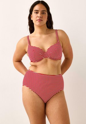 Donna con capelli lunghi e scuri che indossa un bikini a vita alta a righe rosse e bianche, posando contro uno sfondo chiaro e semplice.