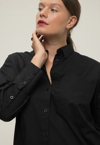 Chemise noire à col, avec une poche poitrine unique et des manches longues avec des poignets à bouton ; texture lisse et coupe ajustée.