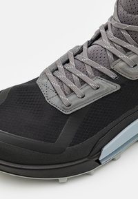 Svart treningstype med mesh-overdel, grå snører, en teksturert design og en fleksibel såle. Har en lys grå aksent på siden.