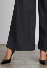 Pantalones de pierna ancha negros con una textura suave, que presentan un dobladillo limpio y un acento en la costura lateral, combinados con zapatos de tacón alto negros y brillantes.