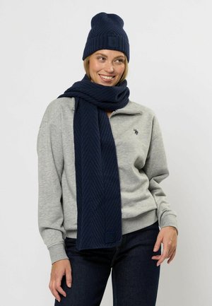 Set de căciulă și eșarfă tricotate, de culoare bleumarin, cu textură ribbed și model herringbone, asortat cu un hanorac gri deschis și blugi închiși la culoare.
