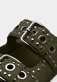 Πράσινο σανδάλι από καστόρι με δύο φαρδιές λουρίδες, ασημένιες στρογγυλές τρουκς και μια μεγάλη μεταλλική πόρπη, με υφή επιφάνειας και ανοιχτό σχέδιο.