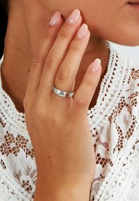 Hand med glänsande ljusrosa naglar och silverring på ringfingret, som rör vid kinden, iklädd en vit spetsblus.