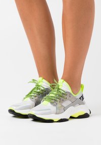 Zapatillas deportivas con una parte superior de malla que presenta tonos plateados y grises reflectantes, acentos en verde neón y suelas de goma negra texturizada.