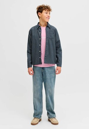 Teen boy standing, wearing a black-and-white checkered shirt over a pink T-shirt, loose blue jeans, and beige shoes, looking to the side.