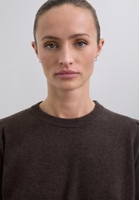 Femeie cu piele deschisă și ochi verzi, purtând cercei mici din argint și un pulover maro închis, îndreptându-se spre cameră pe un fundal gri.