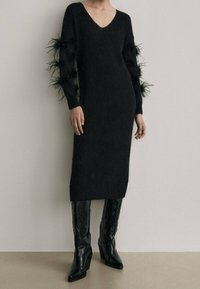 Black knitted dress with a V-neck, featuring feathered accents on long sleeves. Paired with knee-high, black leather heeled boots.