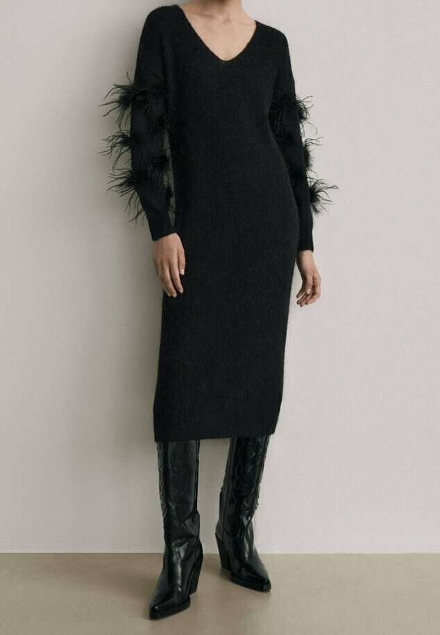 Black knitted dress with a V-neck, featuring feathered accents on long sleeves. Paired with knee-high, black leather heeled boots.