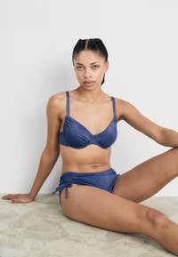 Conjunto de bikini azul marinho com acabamento texturizado. O sutiã tem aro e alças ajustáveis; a parte de baixo é de cintura alta com laços laterais.