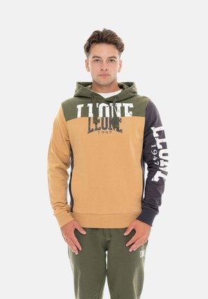 Jonge man draagt een colour-block hoodie met de tekst "LEONE 1947" en bijpassende olijfgroene joggingbroek, staand met handen ontspannen voor zich.