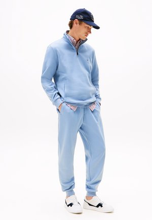 Pullover azzurro chiaro con mezza zip e tasche laterali, abbinato a pantaloni jogger coordinati. Sneakers bianche con accenti neri completano l'outfit.
