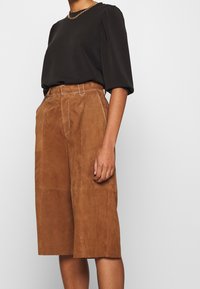 Brown suede culottes with side pockets and prominent stitching details, paired with a black top featuring a round neck and puffed sleeves.