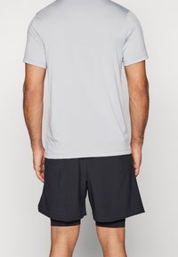 T-shirt de sport gris clair à manches courtes, associé à un short noir. Le matériau semble lisse avec un design ajusté. Détails de couture visibles.