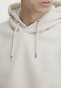 Svetlo béžová mikina s kapucňou a sťahovacou šnúrkou, so striebornými očkami a jemnou textúrou. Má uvoľnený, oversized dizajn.