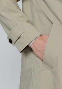 Beige trenchcoat ärm med svart knappdetalj och hand delvis inne i sidofickan mot en plain bakgrund.