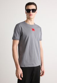 Grå bomulds T-shirt med rund hals og korte ærmer, med et rødt rektangulært logo på brystet. Båret med sorte bukser.