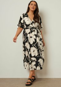 Svart blommig wrapklänning med korta ärmar, har en knytmidja och en lång, flödig silhuett. Blommönster i krämfärger på en mörk bakgrund.
