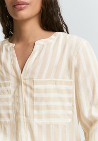 Lys beige skjorte med knapper og hvide vertikale striber, med rund halsudskæring, to forlommer og et struktureret stof.