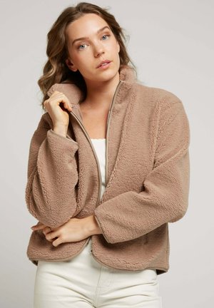 Veste en polaire marron clair avec un col montant, une fermeture éclair et une surface texturée. Présente des manches larges et un ourlet arrondi.